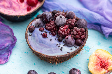 Coconut bowl of frozen raspberries and blackberries on a cyan worn out table top with blue cream. Smoothie bowl with frozen summer fruit.