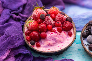 Coconut bowl of frozen strawberries and raspberries with pink cream on a cyan worn out table top. Smoothie bowl of frozen summer fruit.