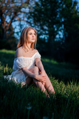 Naklejka na ściany i meble A beautiful young woman is sitting in a field in the green grass. She has a white dress, light hair, bare shoulders, and a happy face. Enjoy nature.