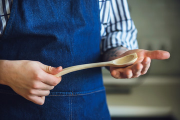 The Woodcarver, dressed in a blue apron and striped shirt, holds a new wooden spoon with an elegant...