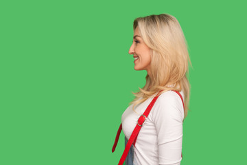 Profile of happy beautiful adult blond woman in stylish overalls looking at copy space with toothy...