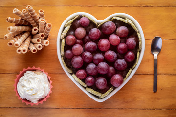 Close-up view from above of white heart-shaped plate full of red grapes, cream and biscuits. Wooden background