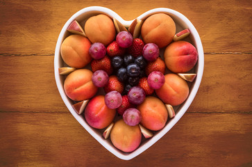 Close-up view from above of a white heart-shaped plate full of red grapes, apricots, strawberry and blueberries. Wooden background