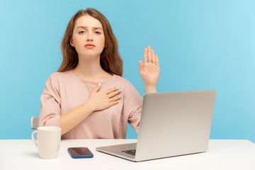 I swear! Responsible honest woman employee sitting at workplace and raising hand to give promise, taking oath with trustworthy faithful expression. indoor studio shot isolated on blue background