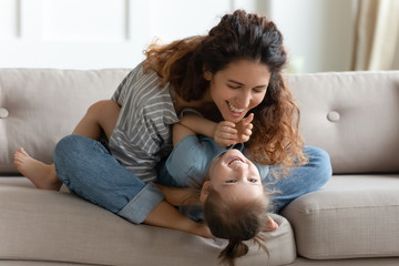 Happy young mother and little daughter having fun, cuddling and playing, sitting on cozy couch at...