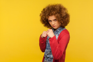Portrait of furious mad woman with curly hair in casual outfit keeping fists up to fight, boxing to defend or attack, looking madly threatening to hit. indoor studio shot isolated on yellow background