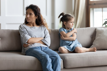 Angry depressed mother and stubborn little daughter ignoring each other after quarrel, pouting, sitting with arms crossed on couch in living room, parent and child, family generations conflict