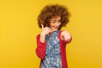 Hey you, call me! Portrait of playful pretty woman with fluffy curly hair showing telephone hand gesture near head, winking and pointing to camera. indoor studio shot isolated on yellow background