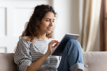 Smiling young woman using computer tablet, sitting on couch, typing on touchscreen, chatting in...