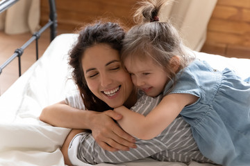 Close up smiling mother and little daughter cuddling in bed, preschool girl lying on happy mum back, hugging, family enjoying tender moment, leisure time, having fun on weekend in bedroom