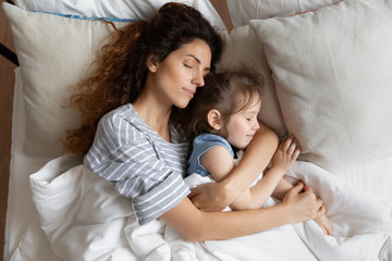Fototapeta na wymiar Top view close up peaceful loving mother and little daughter sleeping on soft pillow together, relaxing lying in cozy bed under warm blanket, caring young mum hugging sleepy preschool girl