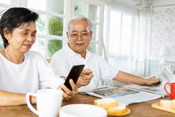 Fototapeta na wymiar Portrait of an elderly Asian couple following the daily news by reading a newspaper and smartphone while eating breakfast. They enjoy and smiling faces. The activity of retirement age