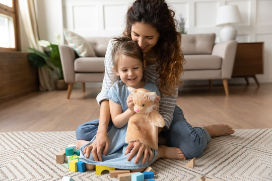 Smiling mother and little daughter hugging, playing with toys, sitting on warm floor with underfloor heating in living room, happy young mum and preschool girl enjoying leisure time together
