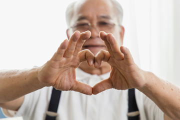Obraz na płótnie Canvas Portrait of an elderly Asian man with a healthy smile and holding a heart-shaped hand near the eyes showing love. Close-up of Retirement health care concept