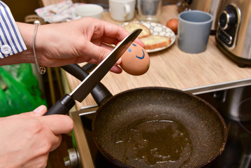woman in the kitchen in front of the pan holds a knife and breaks a smiling egg