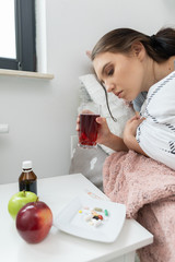 A sick teenager is drinking hot tea with herbs while lying in her bed.