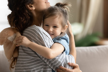 Close up loving mother hugging smiling adorable little daughter, expressing care and protection, cute preschool girl holding fluffy toy, embracing mum, family enjoying tender moment, sitting on couch