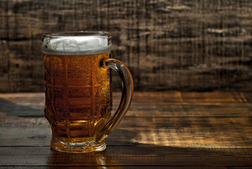 A glass of amber beer. Glasses on a wooden table. Black shabby boards on the background. Bright contrasting light and glare on the glass. Place for text near the booze.