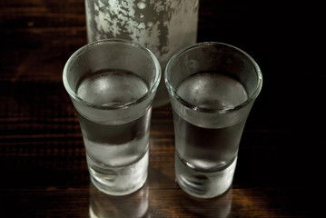 Frozen glasses with alcohol near the bottle. Drinks on a dark brown table. Transparent utensils on a black background. Old shabby boards under glass.