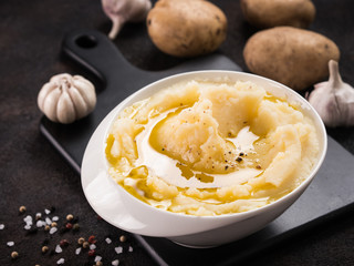 Mashed potatoes with spices and olive oil in white bowl on black cutting board over dark concrete background