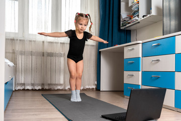 a little girl in a black gymnastics leotard is doing gymnastics at home online in front of a laptop. distance rhythmic gymnastics for children