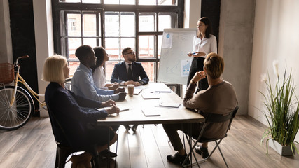 Confident businesswoman coach presenting project statistics on flip chart at corporate meeting in boardroom, mentor leader explaining strategy to diverse employees, training staff at workshop