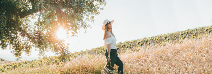 Panoramic of Young woman with a straw hat and a basket walking through the field at sunset in summer. Concept of freedom, tranquility, nature, ecology, green world and happiness. © Martley