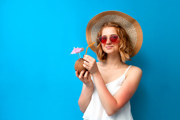 girl with coconut cocktail in a sun hat and glasses on a blue background, a woman with an exotic drink on vacation, summer concept