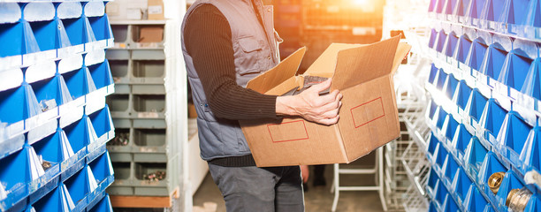 Delivery man in gray uniform holds a filled order box in a warehouse. Golden backlight, widescreen...