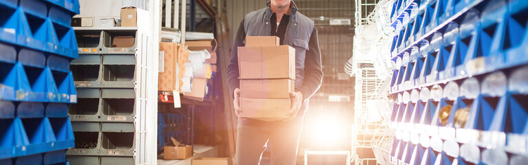 Widescreen image, delivery man in gray uniform carries boxes in his hands at the warehouse. Gold...