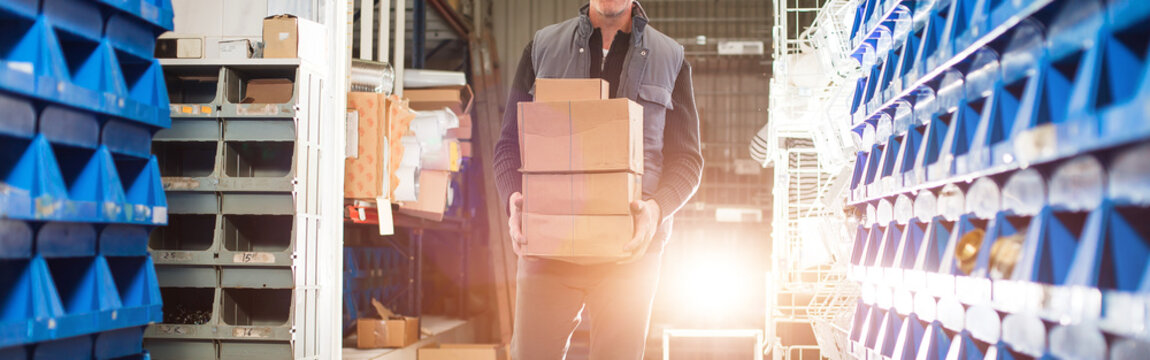 Widescreen image, delivery man in gray uniform carries boxes in his hands at the warehouse. Gold backlight.