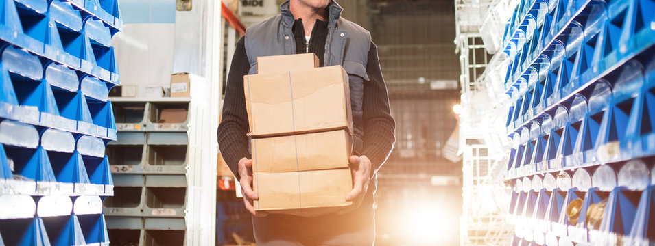 Delivery man in gray uniform holds boxes in his hands at the warehouse. Gold backlight, widescreen image.