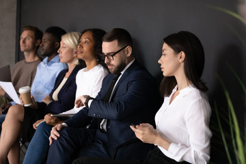 Bored diverse people candidates waiting for job interview, sitting in row in queue in office room,...