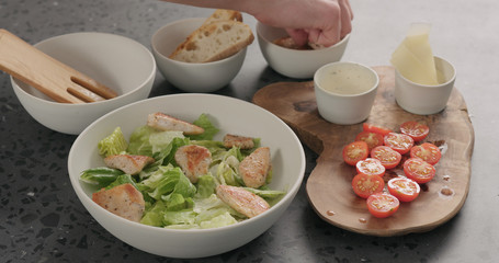Man hand add cherry tomatoes and chicken on caesar salad in white ceramic bowl on concrete countertop
