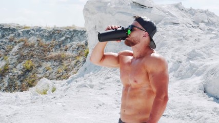 Strong shirtless athlete drinks water from a bottle. Training in sports in nature.
