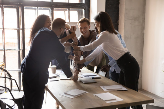 Excited business people stacking fists at table, engaged in team building activity at corporate meeting in boardroom, smiling colleagues celebrating success, teamwork result, business achievement