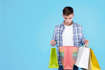 Young man in white t-shirt, checkered shirt is holding colorful paper bags, packages on blue background. Smiling guy student is looking at purchase in packet. Shopping and sale concept. Black friday.