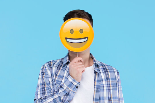 Young Man In White T-shirt, Checkered Shirt Is Holding Paper Decorative Emoticon, Emoji Icon, Sign In Front Of Face. Guy Student Is Posing With Photo Props On Blue Background. Emotional Portrait.