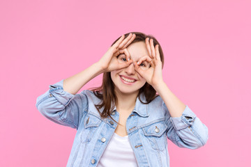 Cheerful smiling girl dressed in white t-shirt, denim jacket on pink background. Young woman is grimacing, showing glasses, mask with hands. Female gesturing. Emotional portrait concept.