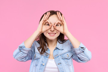 Cheerful smiling girl dressed in white t-shirt, denim jacket on pink background. Young woman is grimacing, showing glasses, mask with hands. Female gesturing. Emotional portrait concept.