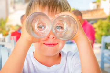 Funny kid looking through two drink glasses as if it were binoculars. Child having fun while waiting for order at cafe, restaurant. Happiness and positive emotions background. Traveling concept