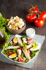 Fresh Caesar salad made of tomato, ruccola, chicken breast, eggs, arugula, crackers and spices. Organic ingredients in a white, transparent bowl on wooden background
