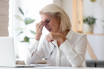 Exhausted mature female employee sit at desk take off glasses massage eyes suffer from headache...