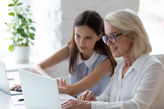 Diverse female coworkers sit at desk in office work together in modern workplace, confident young woman worker help middle-aged colleague with laptop problem or bug, teamwork concept
