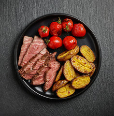 Grilled beef fillet steak with potatoes and tomatoes on black plate on black background, top view