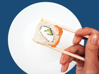 Female hand takes chopsticks sushi rolls with a white round plate on a blue background.