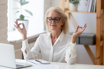 Calm middle-aged businesswoman sit at desk in office relax practice yoga at workplace, peaceful...