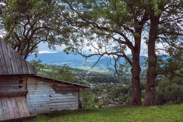 An old wooden house is located on a hill. The village and mountains are further away.