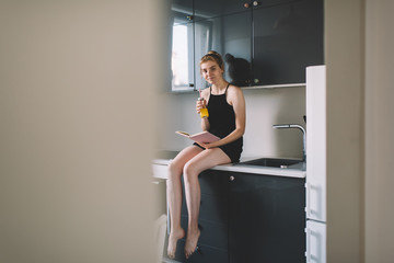 Cheerful female with bottle of drink and book sitting on kitchen counter