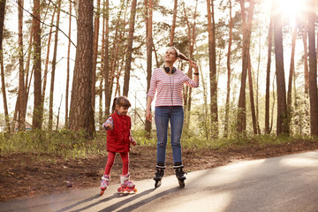 Picture of tender sincere mother standing on road with her daughter, chilling out together, rollerblading, mom teaching her child, enjoying nature being in forest, being on good terms. Family concept.